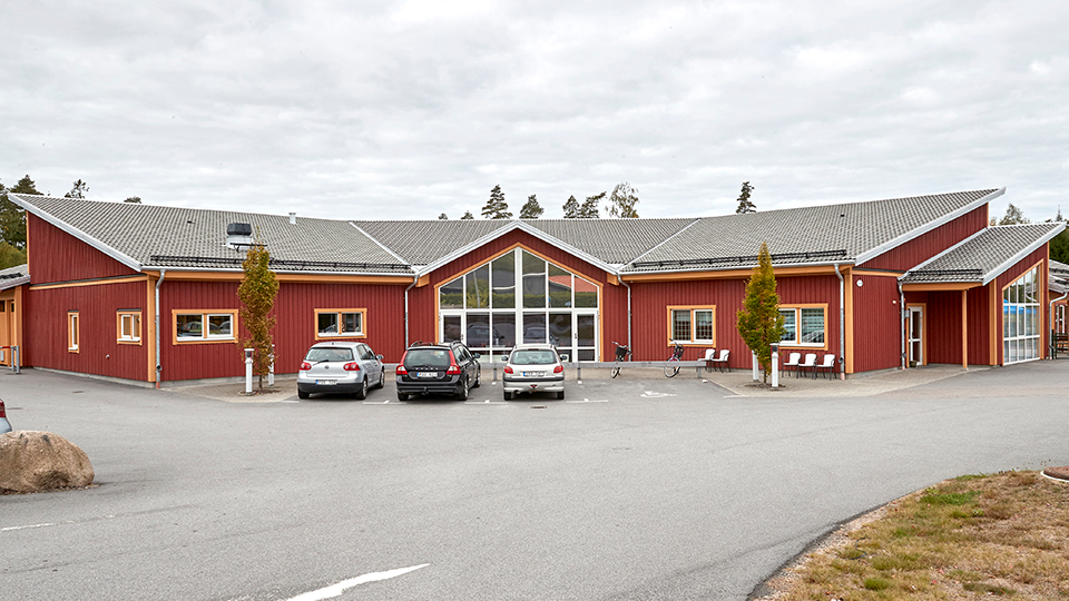 Ett stort rött trähus med grått tak. Framför skolan finns en stor parkering där tre bilar står parkerade.