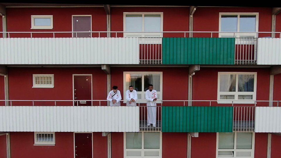 Foto på ett rött lägenhetshus och tre män i vit klädsel som tittar in i kameran.