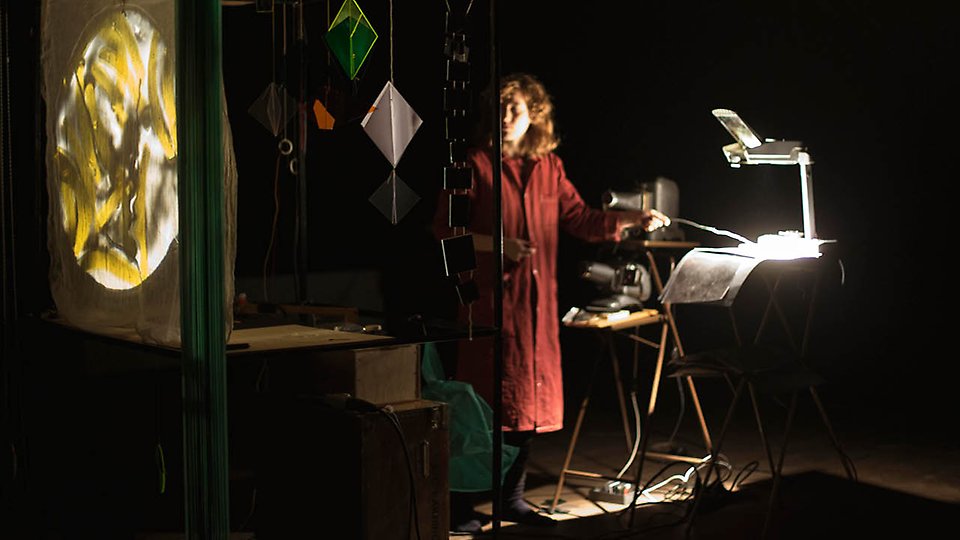 Foto på konstnären Oona Libens i ett mörkt rum, där hon håller objekt framför sina projektorer.