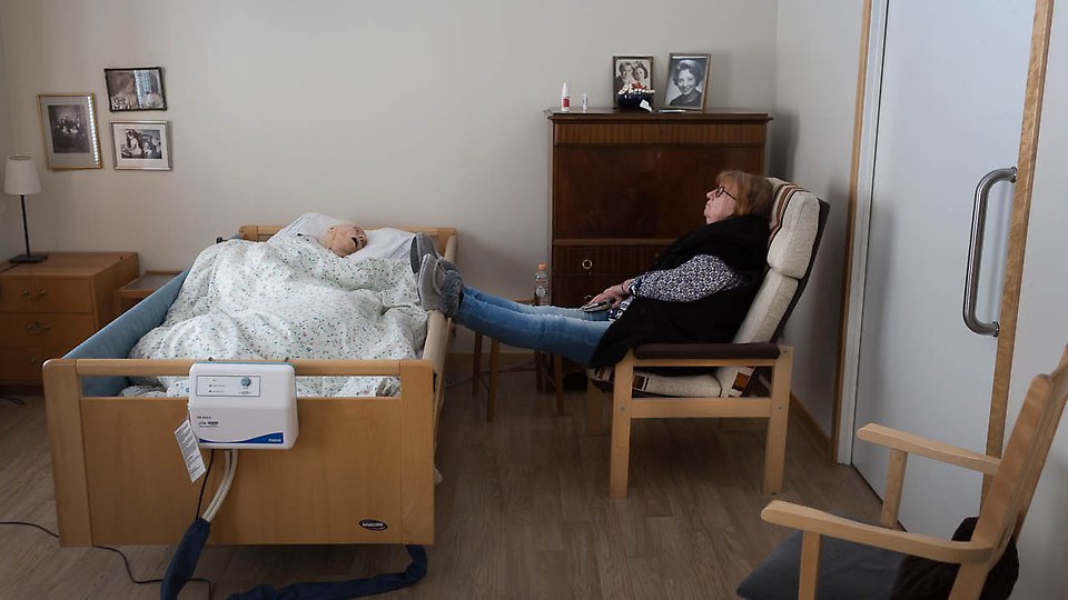 Foto på en gammal person i en sjuksäng, bredvid sitter en anhörig.