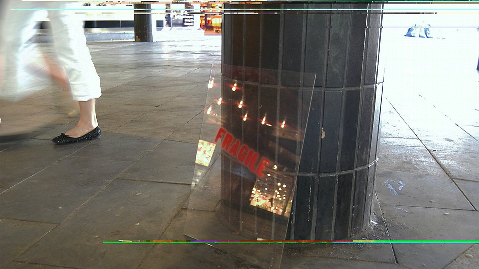 Ett foto på ett torg, där en glasruta står lutad mot en pelare med texten Fragile på. 